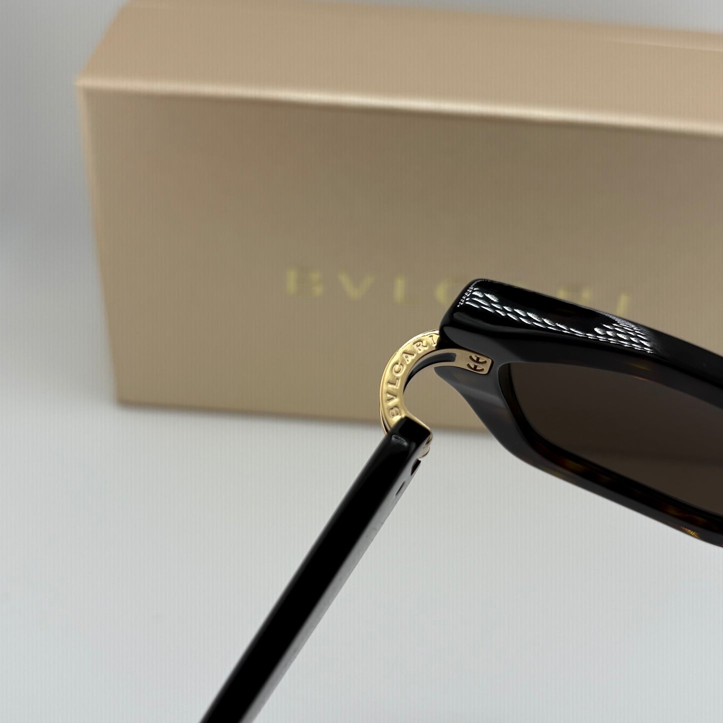 Close-up of a pair of black sunglasses with gold-tone metal details and a patterned temple, shown with a beige Bvlgari box in the background.