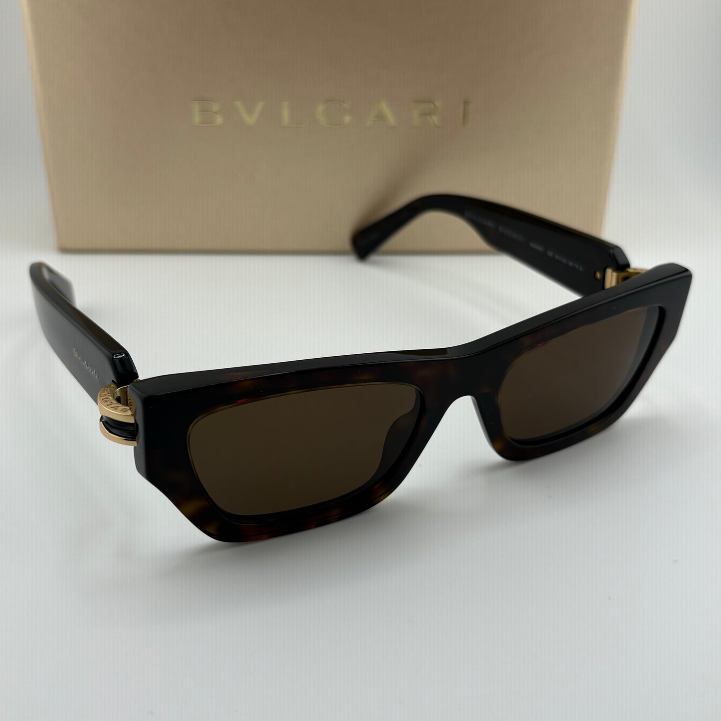 Bvlgari sunglasses with a dark tortoiseshell acetate frame, dark brown lenses, and gold-tone metal accents, shown with their beige Bvlgari branded box in the background.